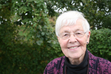 A portrait of a senior, light skinned woman with short white hair wearing thin glasses, a black turtleneck, and a purple sweater. The woman is smiling and standing outside in front of greenery.
