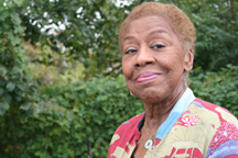 A portrait of a senior medium-dark skinned woman wearing a bright patterned shirt. The woman is smiling and standing outside in front of greenery.