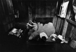 A young boy sits in a small room with wood walls, dressing himself on the side of a bed holding a handful of other children with one blanket over them. To the left, two children lay in a crib.