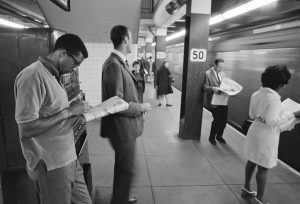 Six people in business attire, spread across the background and foreground, stand on a platform as a train rushes by. The pillars that recede into the background read "50."