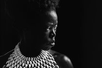 A close up of a heavily-shadowed woman wearing a top made of cowrie shells looks to her left, where he face is illuminated by a light source outside of the frame.