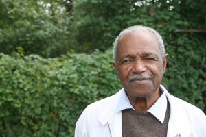 A portrait of a dark skinned man with a mustache wearing a white suit jacket over a brown sweater vest and white collared shirt. The man is softly smiling and standing outside in front of greenery.