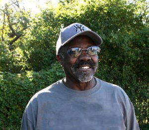 A portrait of a dark skinned man with a mustache and beard wearing a blue baseball cap, tinted glasses, and a blue shirt. The man is smiling, looking into the distance, and standing outside in front of greenery.