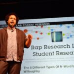 Tahir Hemphill on stage at Creative Time Summit The Curriculum NYC 2015 Brooklyn, NYC. Behind the artist is a slide with the words “Rap Research Lab: Student Research” and the subtitle “The 8 Different Types of N-Word by Gabriel Willoughby."