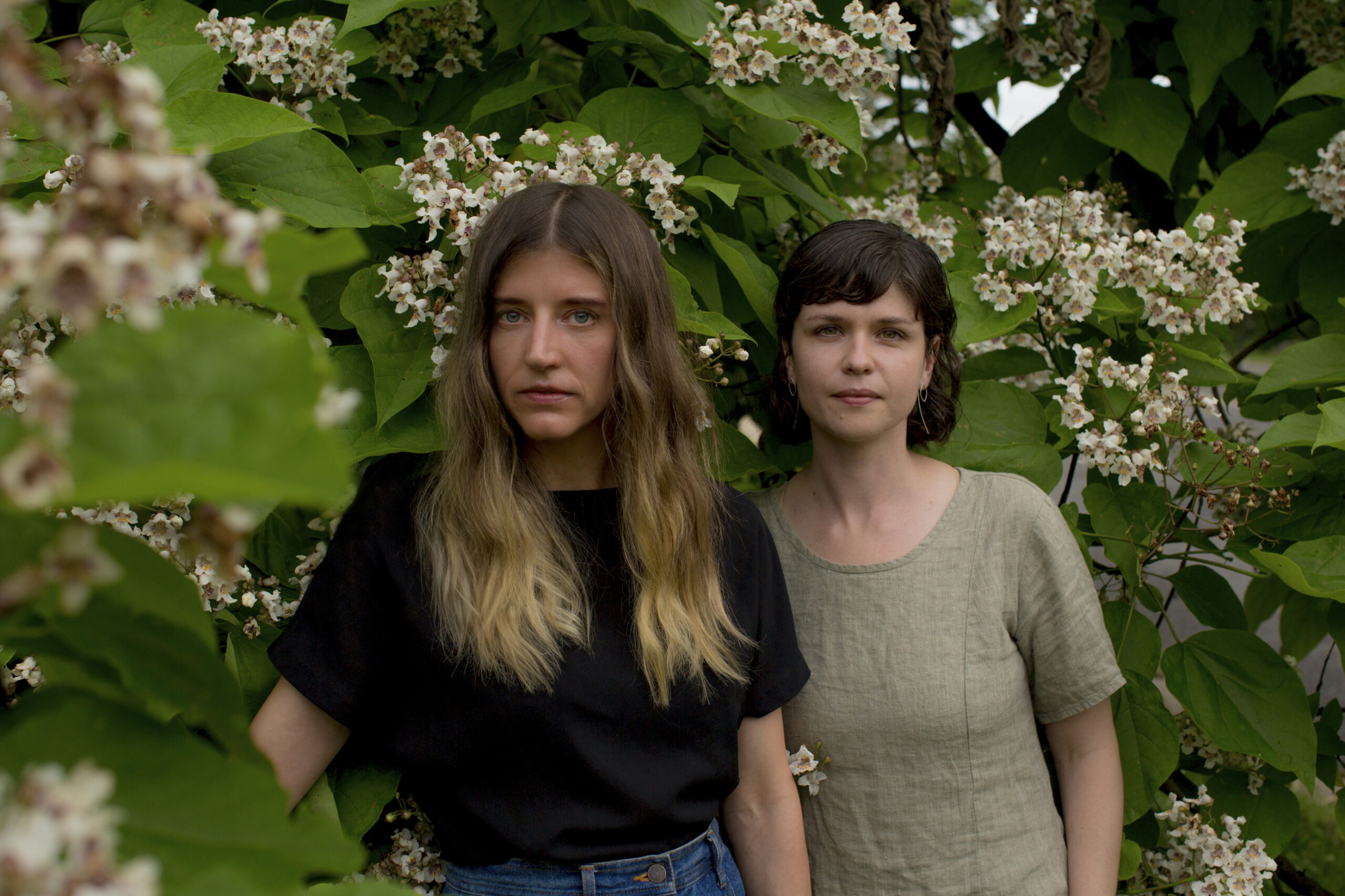 Shannon Collis & Liz Donadio face forward. Collis, on the left, wears a black shirt and has long brown to blond hair. On the right, Donadio wears beige and has dark short hair. Both have light skin tones and stand in front of a large flowering plant.