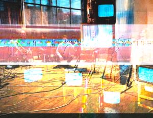 Highly abstracted and layered images include several glowing blue TV monitors. On the left is a window looking out to an urban skyline silhouetted against a blue sky. Wires are scattered across a hardwood floor in the foreground, and translucent figures of seated musicians can be seen superimposed upon the interior space.